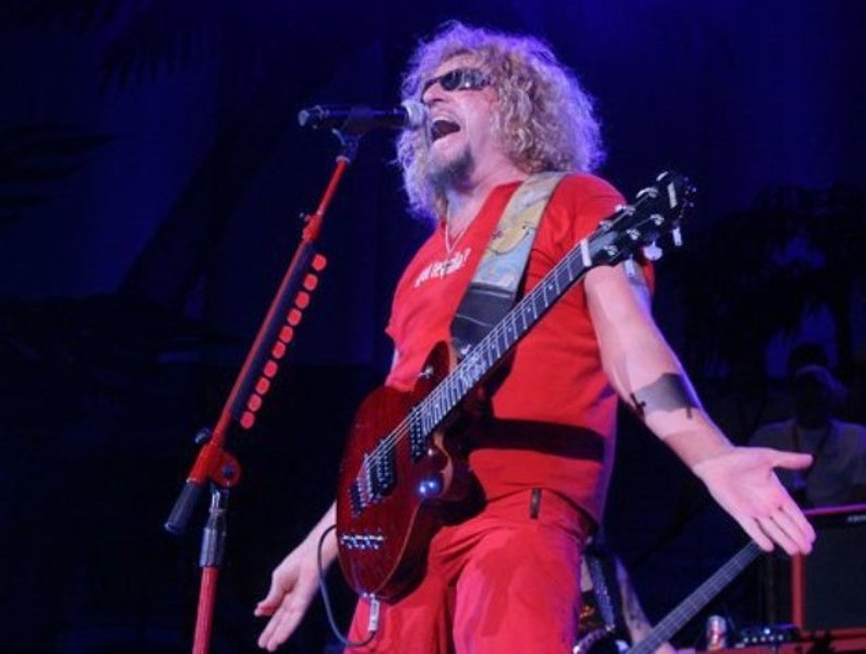Puede incluir: Un hombre con el pelo largo y rubio y gafas de sol canta en un micr&oacute;fono mientras toca una guitarra el&eacute;ctrica roja. Lleva una camisa roja y pantalones rojos.