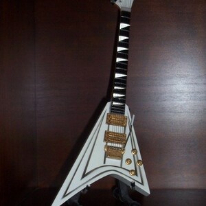 Puede incluir: Una guitarra el&eacute;ctrica blanca en miniatura con cuerpo en forma de V y m&aacute;stil blanco y negro. Cuenta con herrajes dorados y se exhibe sobre un soporte negro. Es una r&eacute;plica.