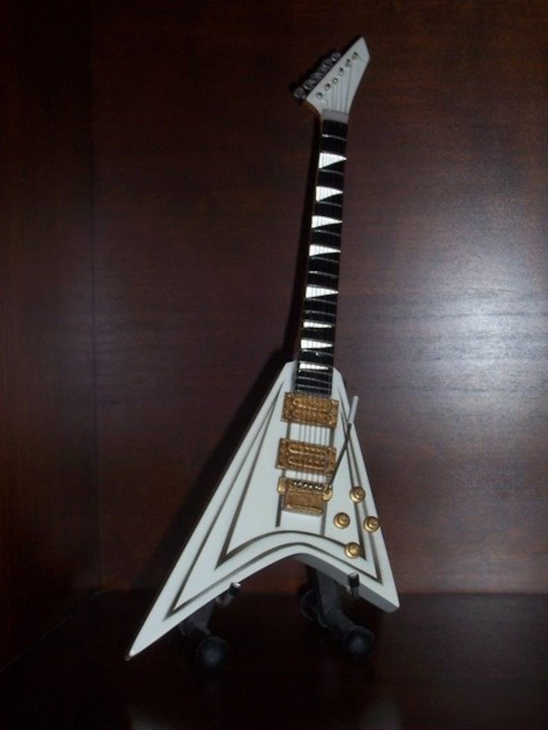 Puede incluir: Una guitarra el&eacute;ctrica blanca en miniatura con cuerpo en forma de V y m&aacute;stil blanco y negro. Cuenta con herrajes dorados y se exhibe sobre un soporte negro. Es una r&eacute;plica.