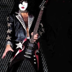 Puede incluir: Un m&uacute;sico con cabello negro y maquillaje est&aacute; tocando una guitarra el&eacute;ctrica negra con detalles rojos. La guitarra tiene una estrella blanca en el cuerpo. El m&uacute;sico lleva una chaqueta de cuero negra con tachuelas plateadas y una estrella blanca en la manga.