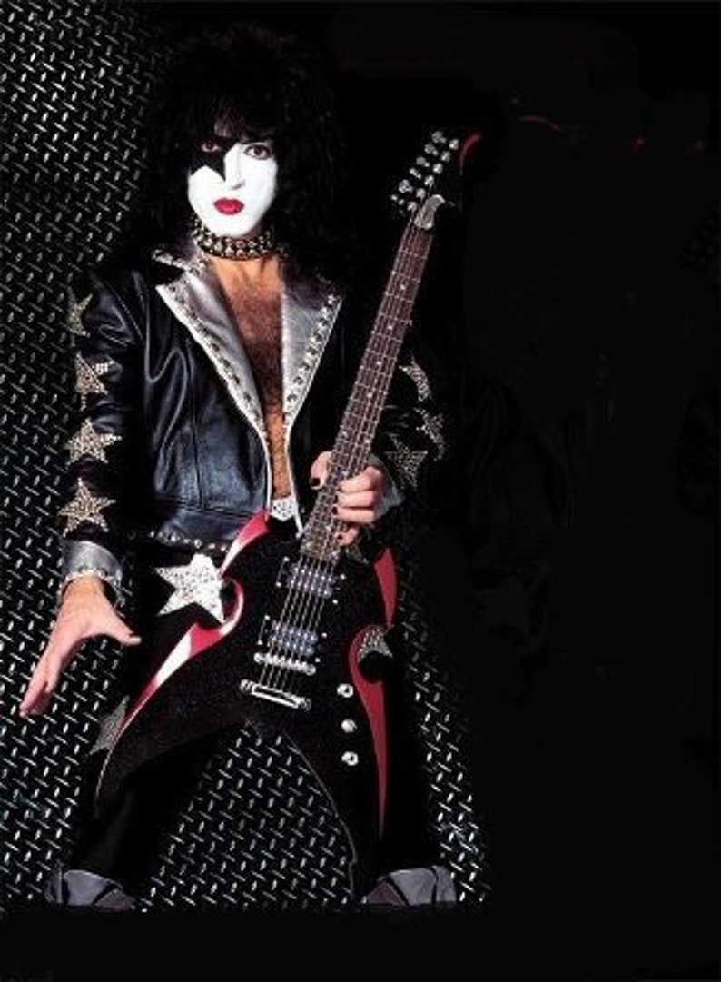 Puede incluir: Un m&uacute;sico con cabello negro y maquillaje est&aacute; tocando una guitarra el&eacute;ctrica negra con detalles rojos. La guitarra tiene una estrella blanca en el cuerpo. El m&uacute;sico lleva una chaqueta de cuero negra con tachuelas plateadas y una estrella blanca en la manga.