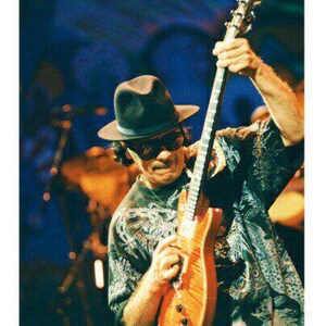 May include: A man wearing a black hat and a patterned shirt plays a brown electric guitar with a black pickguard.