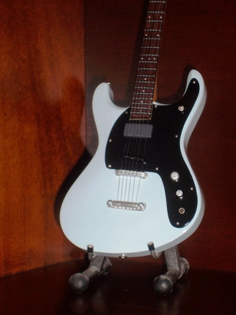 Puede incluir: Una guitarra el&eacute;ctrica azul claro con una golpeador negro y un soporte negro y blanco. La guitarra tiene un solo corte y una forma de cuerpo redondeada.