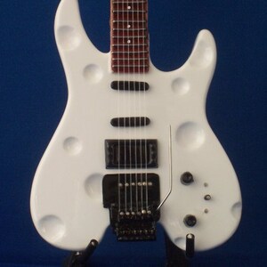 Puede incluir: Una guitarra el&eacute;ctrica blanca con un dise&ntilde;o de cuerpo &uacute;nico y contorneado. La guitarra tiene una golpeador negro y un puente negro. La guitarra est&aacute; en un soporte.