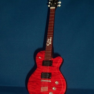 Puede incluir: Una guitarra el&eacute;ctrica roja con una golpeador negro y herrajes cromados. La guitarra tiene un logotipo "Cibson Vibro" en la pala.