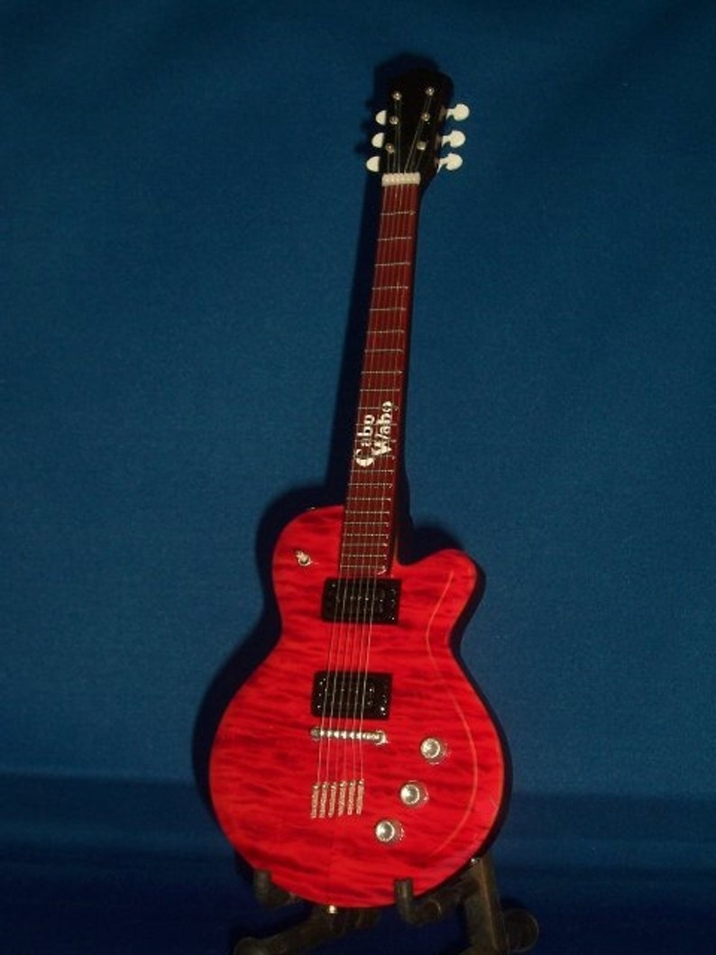 Puede incluir: Una guitarra el&eacute;ctrica roja con una golpeador negro y herrajes cromados. La guitarra tiene un logotipo "Cibson Vibro" en la pala.