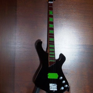 Puede incluir: Una guitarra el&eacute;ctrica de bajo negra con una franja verde a lo largo del m&aacute;stil. La guitarra est&aacute; en un soporte negro.