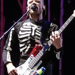 May include: A musician wearing a black and white skeleton shirt and black pants plays a white and blue electric bass guitar with colorful stickers and paint splatters. The bass guitar has a red, white, and blue pickguard.