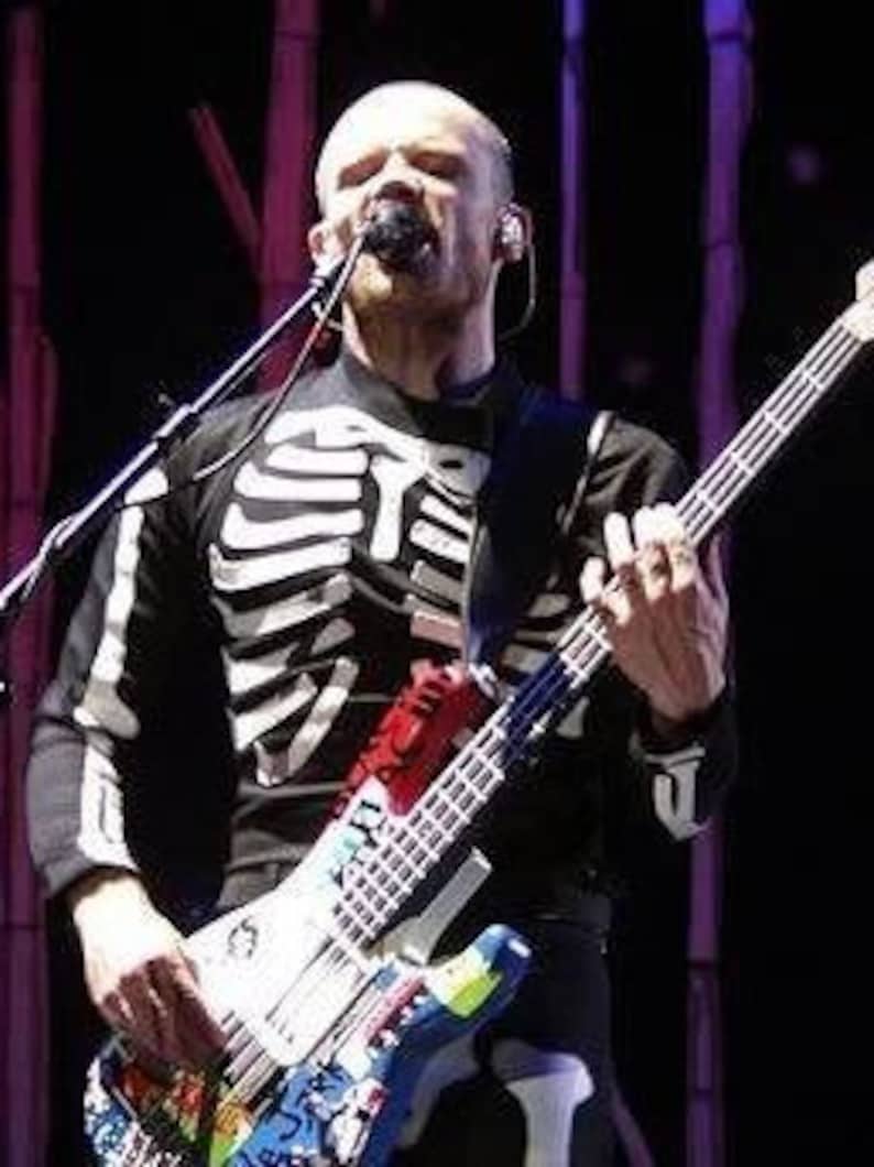 May include: A musician wearing a black and white skeleton shirt and black pants plays a white and blue electric bass guitar with colorful stickers and paint splatters. The bass guitar has a red, white, and blue pickguard.