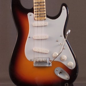 May include: A sunburst finish electric guitar with a white pickguard and chrome hardware. The guitar has a maple neck and a rosewood fretboard.