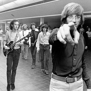 May include: A group of people, some holding guitars, stand in a hallway. One person is pointing directly at the camera.