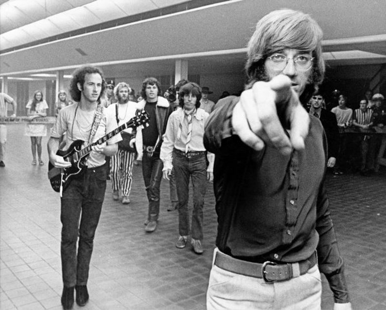 May include: A group of people, some holding guitars, stand in a hallway. One person is pointing directly at the camera.