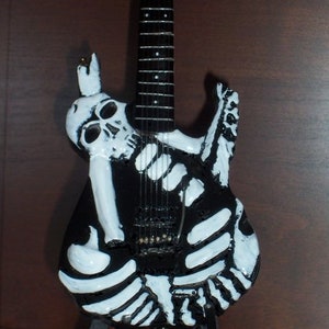 May include: A black electric guitar with a white skeleton design. The guitar has a black body and a black neck with white fret markers. The skeleton design is painted on the body of the guitar.