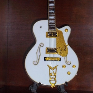 May include: A white electric guitar with gold hardware and a gold inlay on the body. The guitar is on a black stand.