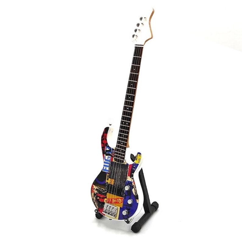 May include: A white electric bass guitar with a multi-colored design featuring various band names and logos, including "Elvis", "Sex Pistols", and "The Clash". The guitar is on a black stand.