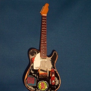 May include: A black electric guitar with a white pickguard and a variety of stickers on the body. The guitar is on a black stand.