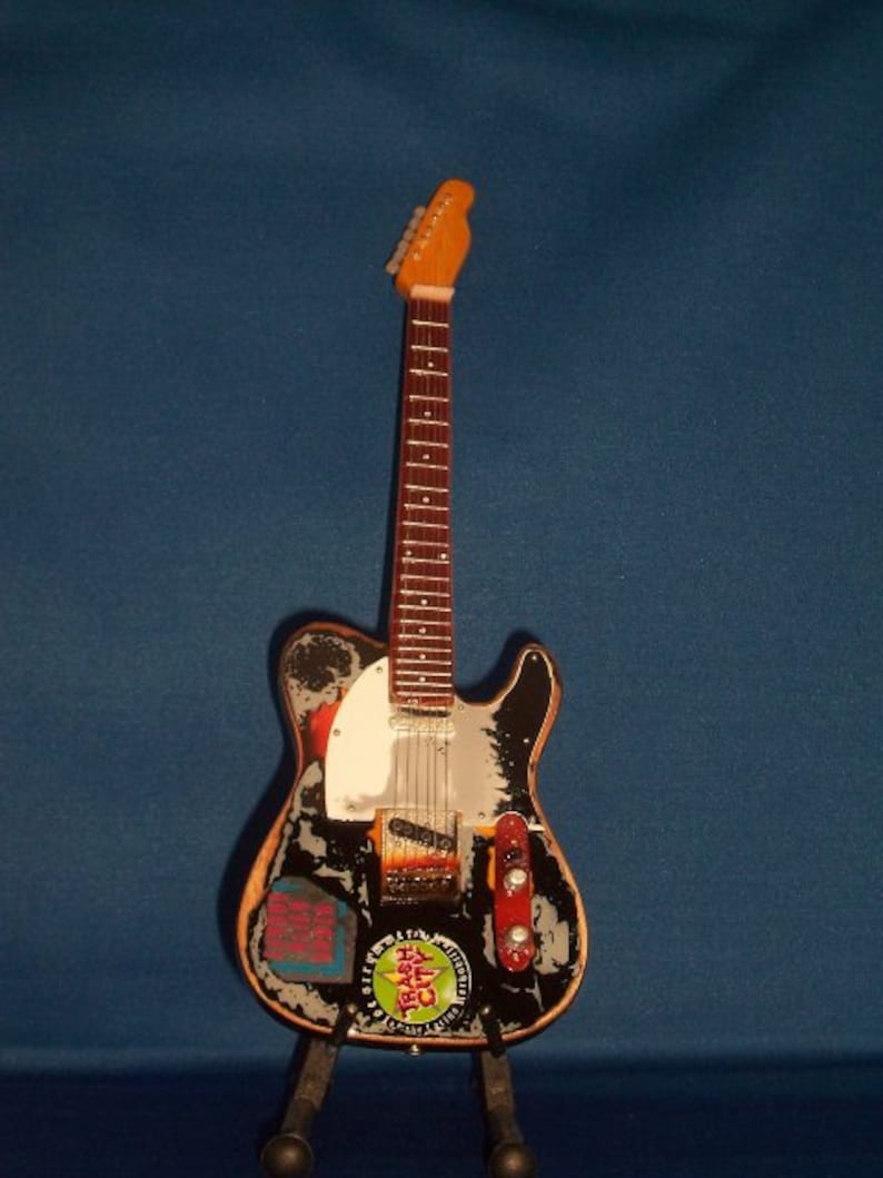 May include: A black electric guitar with a white pickguard and a variety of stickers on the body. The guitar is on a black stand.