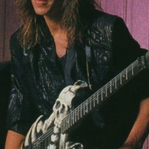 May include: A man with long, dark hair is playing a black electric guitar with a white skull design. The guitar has a black strap and a silver pickguard.