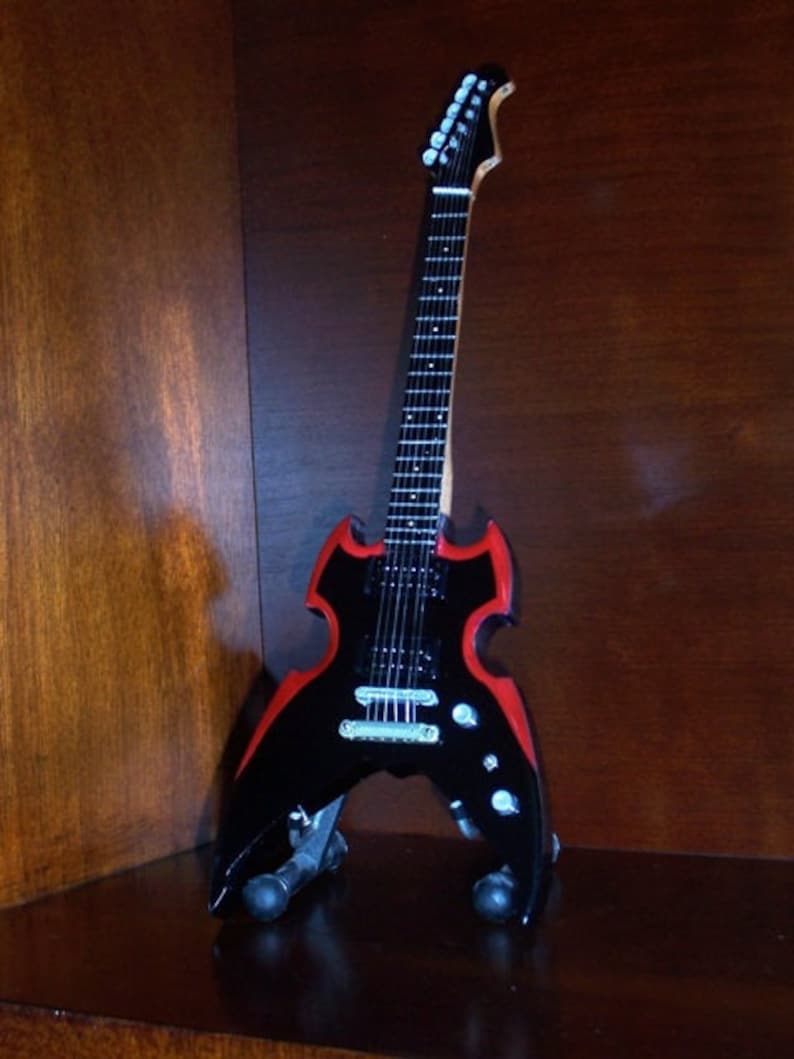 Puede incluir: Una guitarra el&eacute;ctrica negra con un borde rojo, una forma de cuerpo &uacute;nica y una pala plateada. La guitarra est&aacute; en un soporte.