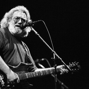 Puede incluir: Imagen en blanco y negro de un m&uacute;sico tocando una guitarra el&eacute;ctrica y cantando a un micr&oacute;fono. La guitarra tiene una forma &uacute;nica y un dise&ntilde;o decorativo. El m&uacute;sico lleva gafas y tiene una larga barba.