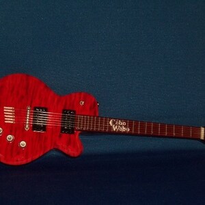 Puede incluir: Una guitarra el&eacute;ctrica roja con una golpeador negro y un logotipo "Cabo Wabo" blanco en el cuello. La guitarra tiene una tapa de arce flameado y un cuerpo de caoba.