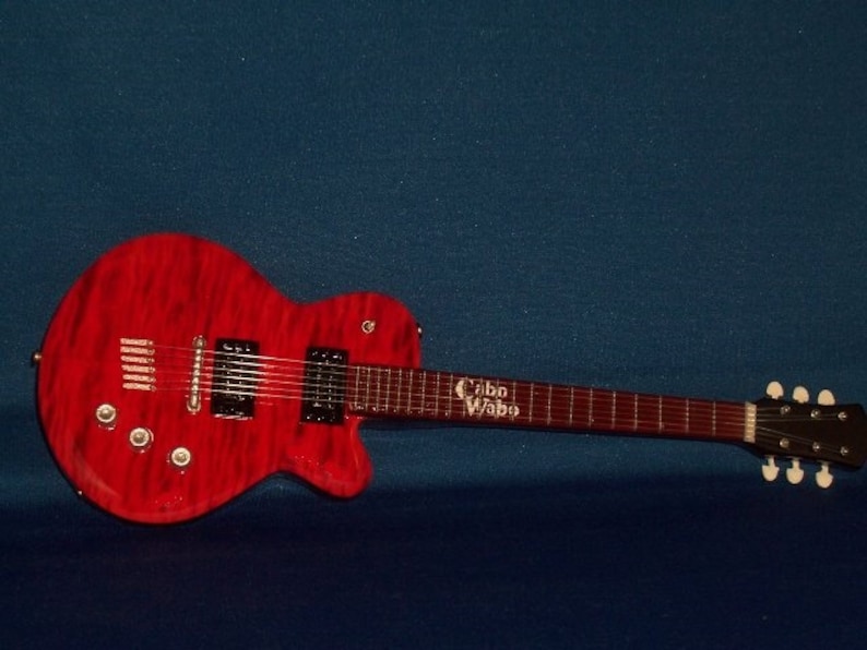 Puede incluir: Una guitarra el&eacute;ctrica roja con una golpeador negro y un logotipo "Cabo Wabo" blanco en el cuello. La guitarra tiene una tapa de arce flameado y un cuerpo de caoba.