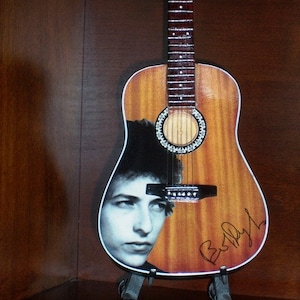 May include: A brown acoustic guitar with a black and white photo of Bob Dylan on the body. The guitar has a black pickguard and a silver signature that says "Bob Dylan".