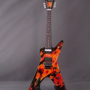 Puede incluir: Una guitarra el&eacute;ctrica negra con un dise&ntilde;o de llamas en naranja y rojo. La guitarra tiene una pala puntiaguda y un cuerpo en forma de V voladora.