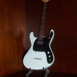 Puede incluir: Una guitarra el&eacute;ctrica azul claro con una golpeador negro y un cuello de madera. La guitarra est&aacute; en un soporte.