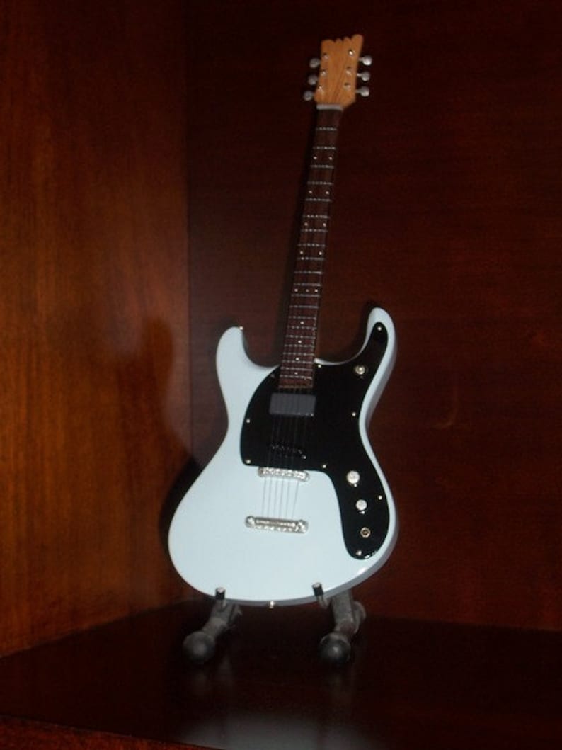Puede incluir: Una guitarra el&eacute;ctrica azul claro con una golpeador negro y un cuello de madera. La guitarra est&aacute; en un soporte.