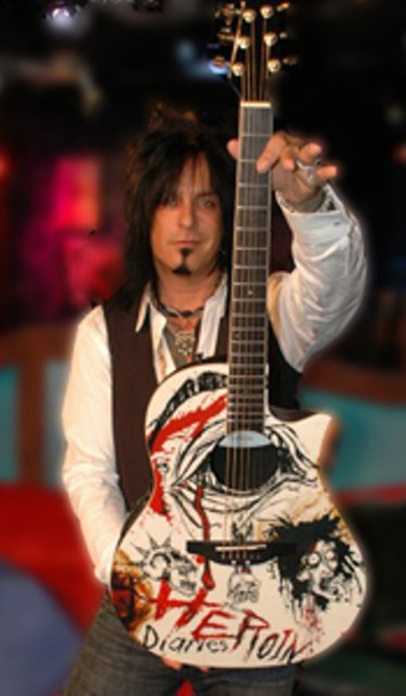 Puede incluir: Un hombre con el pelo largo y negro sostiene una guitarra el&eacute;ctrica blanca con detalles en negro y rojo. La guitarra tiene un dise&ntilde;o de calavera y huesos cruzados con las palabras "The Diaries" y "Rock" escritas en ella.