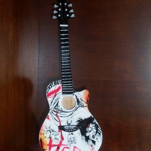 Puede incluir: Una guitarra ac&uacute;stica blanca con dise&ntilde;os gr&aacute;ficos negros y rojos. La guitarra tiene una pala negra con clavijas de afinaci&oacute;n plateadas y un diapas&oacute;n negro. La guitarra est&aacute; en un soporte negro.