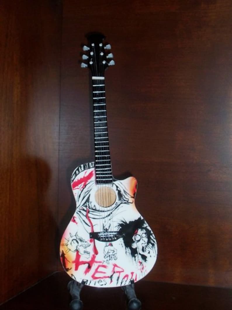 Puede incluir: Una guitarra ac&uacute;stica blanca con dise&ntilde;os gr&aacute;ficos negros y rojos. La guitarra tiene una pala negra con clavijas de afinaci&oacute;n plateadas y un diapas&oacute;n negro. La guitarra est&aacute; en un soporte negro.