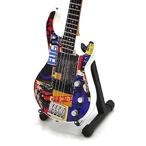 May include: A white electric bass guitar with a multi-colored design featuring band names and logos, including "Joy Division", "Sex Pistols", and "New Order". The guitar is on a black stand.
