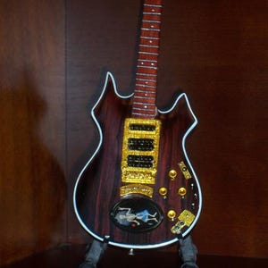 May include: A miniature electric guitar with a dark wood grain finish, white trim, and gold hardware. It features three black pickups, gold control knobs, and a small oval inlay on the body depicting a skeleton figure wearing blue shorts.
