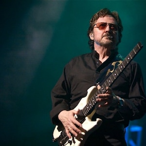 Puede incluir: Un hombre con gafas de sol y una camisa negra toca una guitarra el&eacute;ctrica blanca con un dise&ntilde;o de patr&oacute;n blanco y negro.