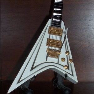 Puede incluir: Una guitarra el&eacute;ctrica blanca con cuerpo en forma de V y herrajes dorados. La guitarra tiene un diapas&oacute;n blanco y negro y se exhibe sobre un soporte negro. El cuerpo est&aacute; acentuado con adornos dorados.