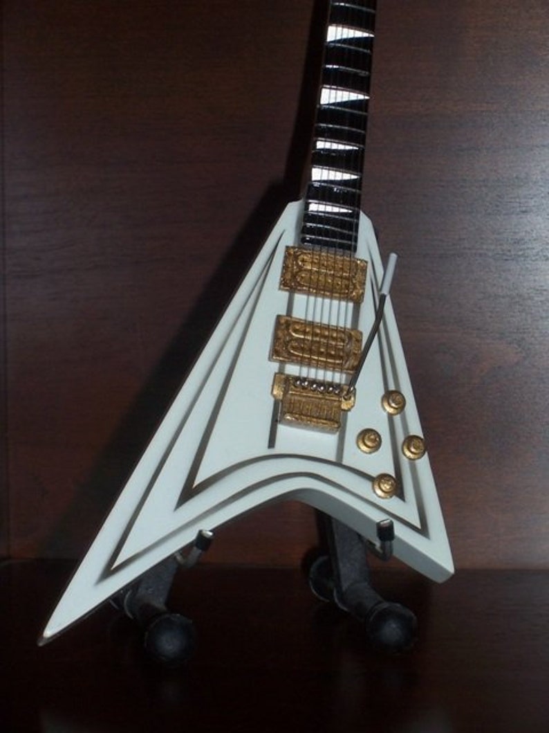 Puede incluir: Una guitarra el&eacute;ctrica blanca con cuerpo en forma de V y herrajes dorados. La guitarra tiene un diapas&oacute;n blanco y negro y se exhibe sobre un soporte negro. El cuerpo est&aacute; acentuado con adornos dorados.