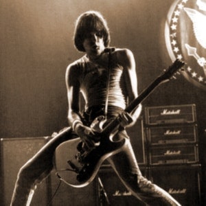 Puede incluir: Una persona tocando una guitarra el&eacute;ctrica negra con una golpeador blanco. La guitarra tiene un solo corte y una correa negra. La persona lleva una camiseta sin mangas blanca y pantalones vaqueros.