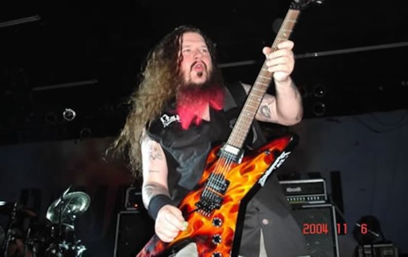 Puede incluir: Un hombre con el pelo largo y rojo rayado toca una guitarra el&eacute;ctrica negra y naranja con llamas pintadas en el cuerpo. La guitarra tiene una golpeador negro y una clavija de afinaci&oacute;n plateada.