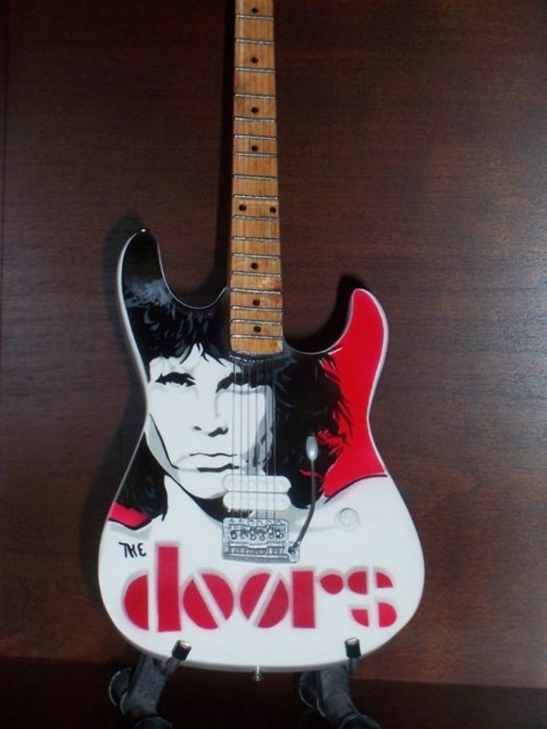 May include: A white electric guitar with a black and white image of Jim Morrison from The Doors band on the body. The guitar has red text that says "The Doors" on the body.