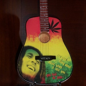 Puede incluir: Una guitarra ac&uacute;stica roja, amarilla y verde con una imagen en blanco y negro de Bob Marley y el texto "One good thing about music, when it hits you, you feel no pain..."