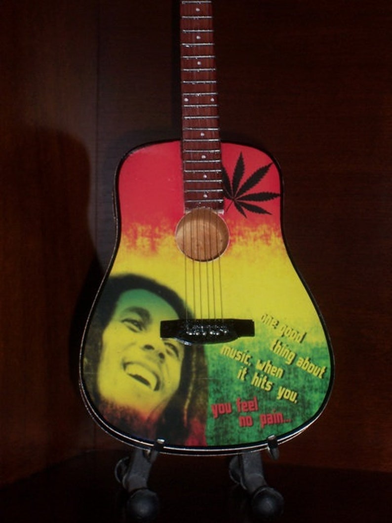Puede incluir: Una guitarra ac&uacute;stica roja, amarilla y verde con una imagen en blanco y negro de Bob Marley y el texto "One good thing about music, when it hits you, you feel no pain..."