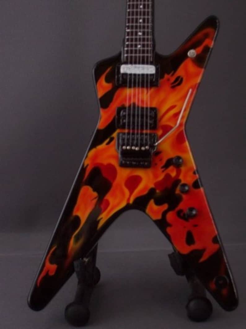 Puede incluir: Una guitarra el&eacute;ctrica negra con un dise&ntilde;o de llamas en naranja y amarillo. La guitarra est&aacute; en un soporte.