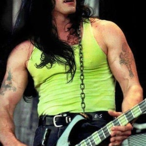 Puede incluir: Un hombre con el pelo largo y negro, que lleva un top sin mangas verde lima y un cintur&oacute;n negro con una hebilla plateada, toca un bajo negro y blanco con una cadena plateada alrededor del cuello.