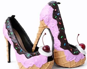 May include: A pair of whimsical high-heeled shoes designed to resemble ice cream cones. The shoes feature a pink ice cream scoop design with chocolate fudge, sprinkles, and a cherry on top. The heels are black.