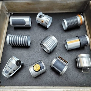 May include: A collection of nine silver and black lightsaber parts, including a hilt, a blade, and a power cell. The parts are arranged in a drawer.