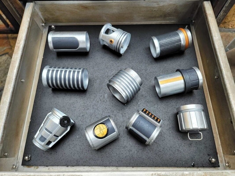 May include: A collection of nine silver and black lightsaber parts, including a hilt, a blade, and a power cell. The parts are arranged in a drawer.