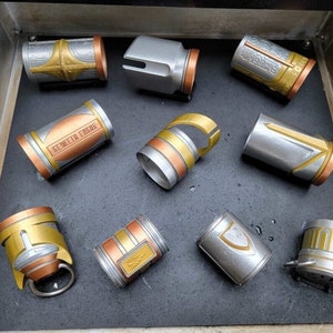 May include: A collection of ten silver and gold lightsaber parts, including a hilt, a coupler, and several different colored sleeves. The sleeves have various designs and markings, including the words "BETWEEN FORCE".
