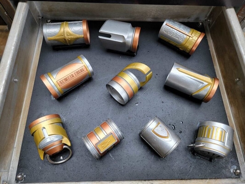 May include: A collection of ten silver and gold lightsaber parts, including a hilt, a coupler, and several different colored sleeves. The sleeves have various designs and markings, including the words "BETWEEN FORCE".